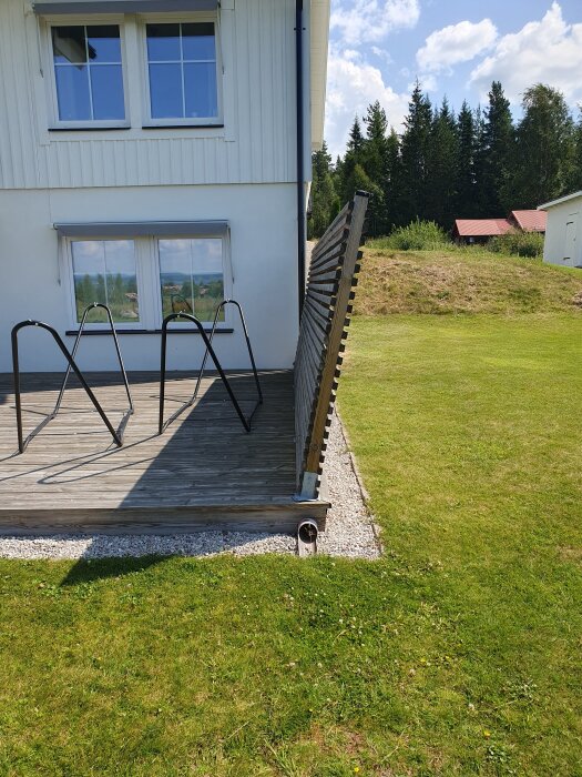 Ett insynsskydd lutar vid sidan av en altan vid ett hus. Gräsmatta och träd i bakgrunden under en klarblå himmel.