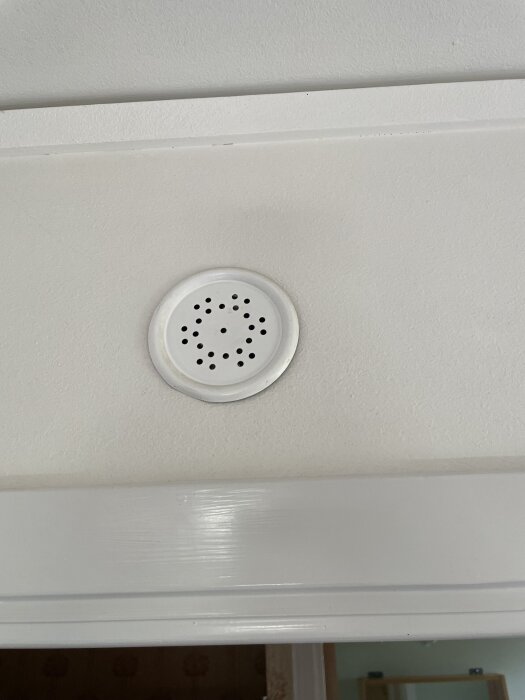 Inbyggd vit ringklocka eller högtalare med hål, monterad i taket ovanför en dörrkarm.