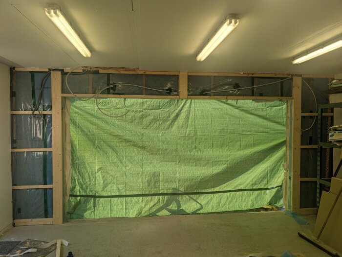 Bilden visar en bredare portöppning inomhus, omgiven av träreglar, med en grön presenning uppspänd där dörren ska monteras.