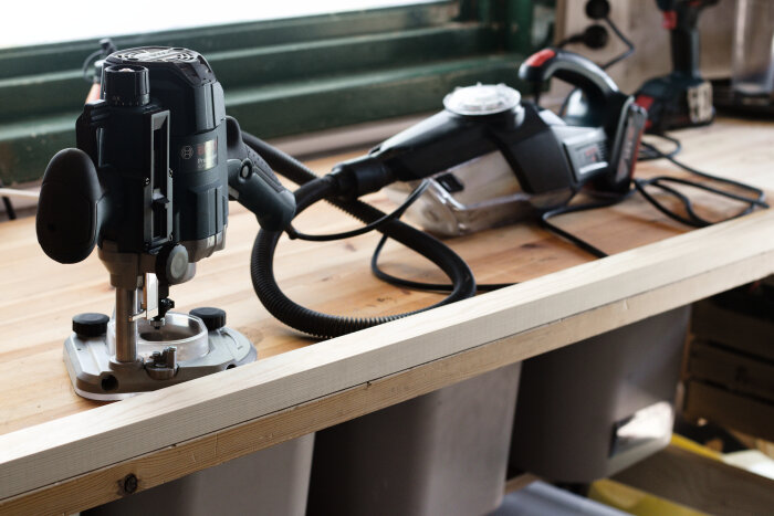 Arbetsbänk med en elektrisk fräs och en dammsugare används för att bygga en skohylla. Verktygen ligger på en träarbetsyta nära ett fönster.