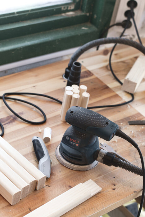 Rundstavar och slipverktyg på en arbetsbänk i trä, verktyg inkluderar en Bosch slipmaskin och en Hultafors Vikbar kniv.