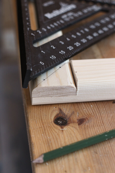 En närbild av trädelar med en urfasning, mätt med en svart vinkelhake; en grön penna syns i förgrunden på arbetsbänken.