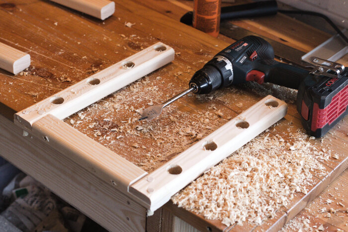 Träkonsoler med borrade hål och sågspån på en arbetsbänk, borrmaskin synlig.