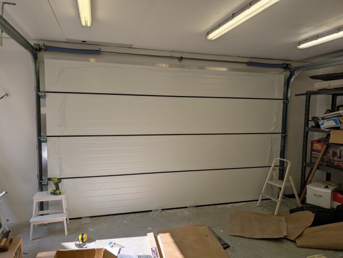 Vit garageport monterad med nya reglar, omgiven av verktyg och stegar inuti ett garage med lysrörsbelysning i taket.