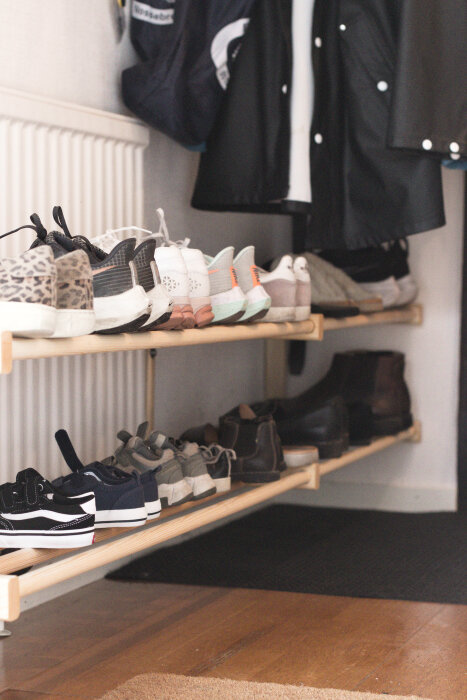 Skohylla i trä med två hyllplan fyllda av olika sneakers och kängor, placerad mot en vägg nära ett element och ytterkläder.