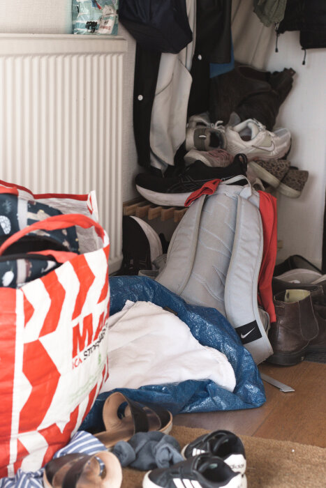 Trång hall med skor, jackor och diverse föremål, inklusive en shoppingkasse och ryggsäck, bredvid ett element.