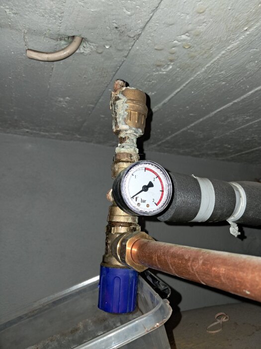 Tryckmätare och ventil på en kopparrörsanslutning, med rost och slitage synligt, samt en blå plastbehållare under i ett grått rum.