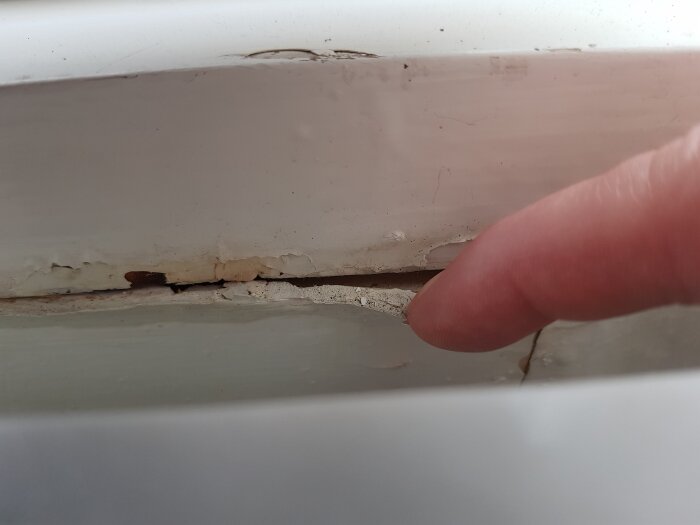 En finger pekar på en spricka i fönsterbänken, vilket visar på skador och bristande underhåll under köksfönstret i en äldre byggnad.