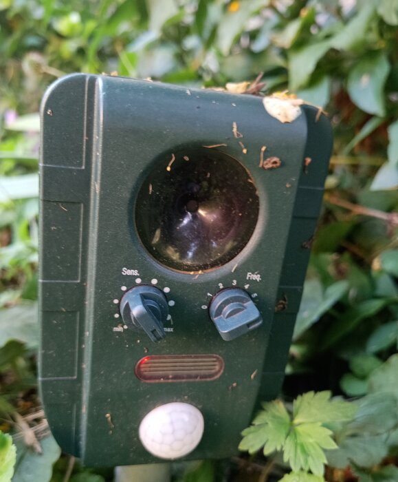 Grön viltskrämmare i trädgårdsmiljö med inställningar för känslighet och frekvens, omgiven av löv och växter.