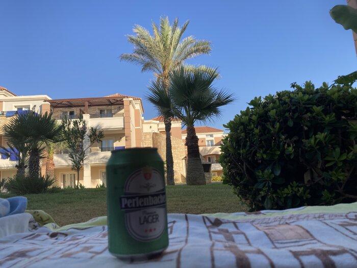 En burk Perlenbacher lager står på en filt, omgiven av palmer och buskar framför en byggnad i solskenet på Kreta.