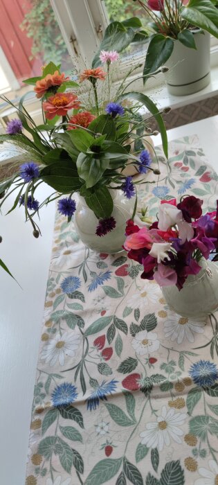Två vaser med buketter av luktärt, zinnia, klint och buketthirs på ett bord med blommigt bordslöpare.