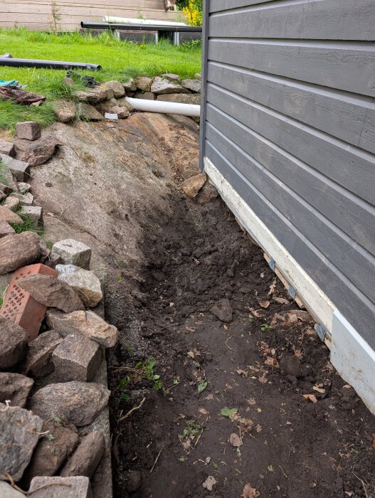 Grop vid attefallshus efter bortgrävd rabatt, synligt berg med stenar runt och ett avloppsrör, nära husets hörn där vatten samlas vid regn.