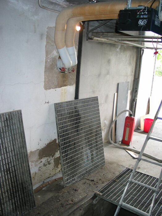 Garage med smörjgrop, två rör i väggen, galler på marken, brandsläckare och stege synliga.