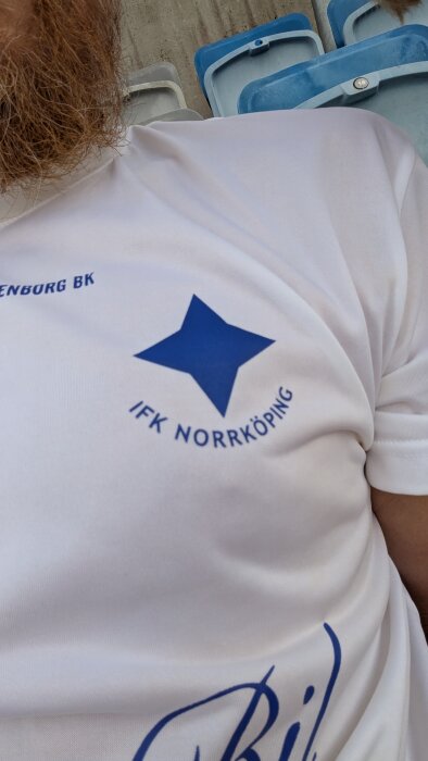 Person i vit t-shirt med blå stjärna och texten "IFK Norrköping" på bröstet, sittandes på en stadionplats med blåa säten i bakgrunden.