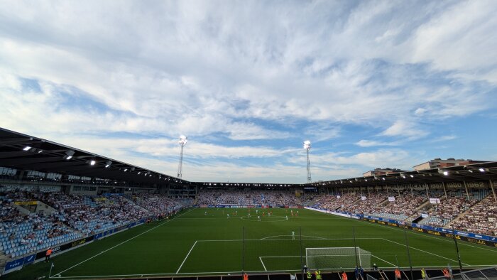 Fotbollsmatch pågår på en stor stadion under en klar himmel, med åskådare som fyller läktarna och strålkastare som lyser upp planen.