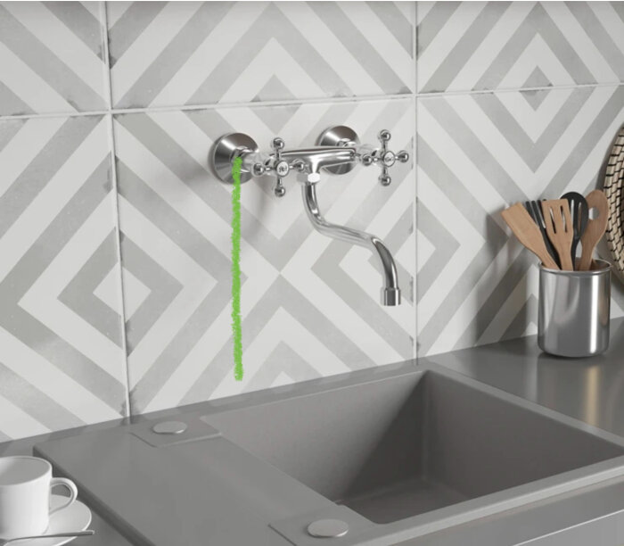 Vägghängd köksblandare över en diskbänk med grönt streck markerat, mot mönstrad kakelvägg.