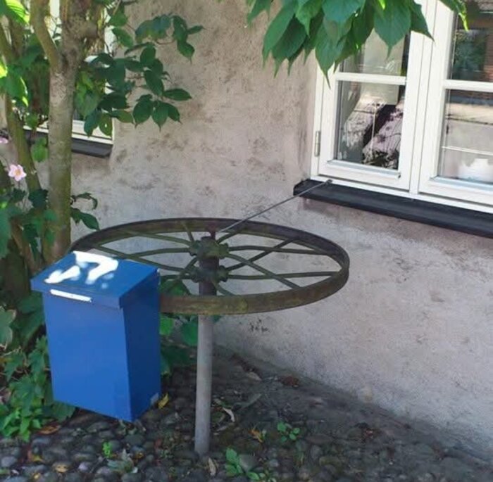 Blå postbrevlåda fäst vid en äldre metallkonstruktion, stående vid ett fönster med en rem eller lina ansluten till huset.