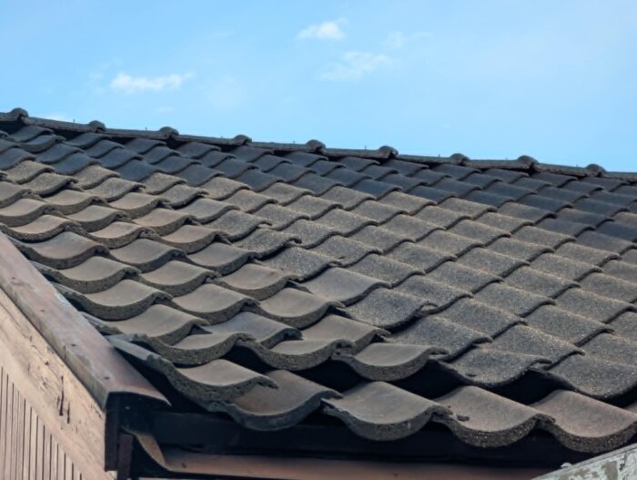 Tak med betongpannor, nedersta raden har en extra panna. Vindskivor och läkten syns, viss färgslitage. Klar himmel ovanför.