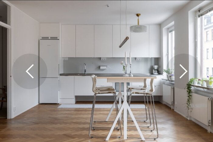 Modernt kök med vita skåp, träbord med konstnärlig bas, höga stolar, ljuskrona och växter i fönsterbrädan.