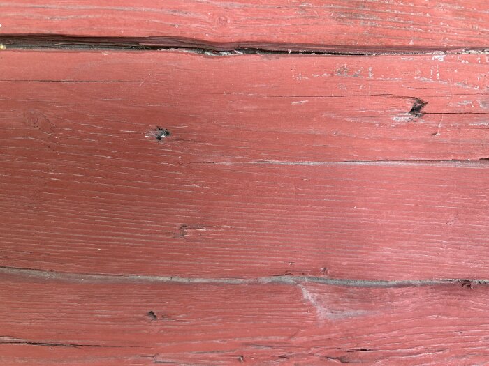 Röd trävägg med synliga sprickor och spikhål, del av ett äldre timmerhus.