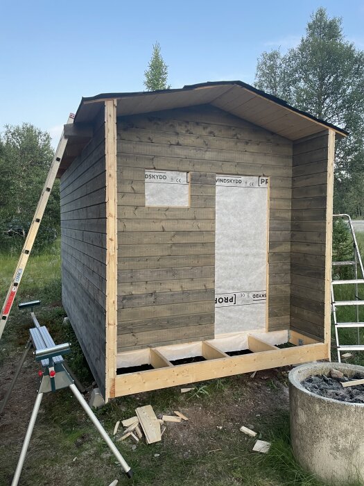 Byggnad under konstruktion med träpanel och skyddspapp, stegar och verktyg synliga i bakgrunden, gräsomgiven byggplats.