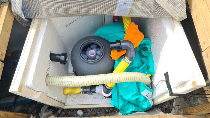 Poolutrustning i tekniklåda med filterbollsfylld behållare, gula och gråa slangar, orange duk och ventiler, placerad på plastduk.