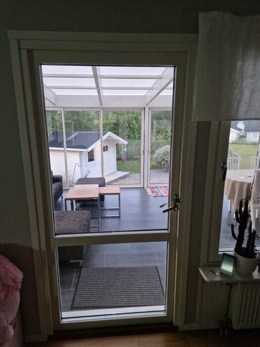 Altandörr med handtag, delvis öppnad, visar glipa i hörnet ovanför handtaget. Utsikt mot inglasad veranda med bord och stolar.