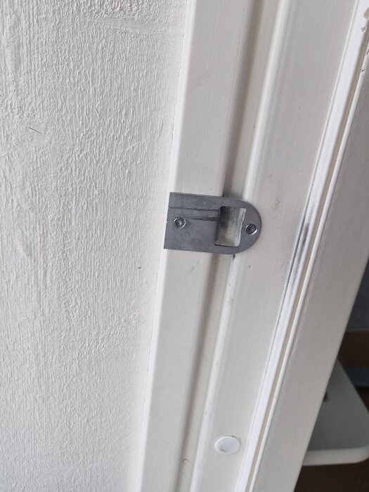 Närbild av en vit dörrkarm med ett metallslutbleck för låsmekanism, visar märken och ojämnheter.