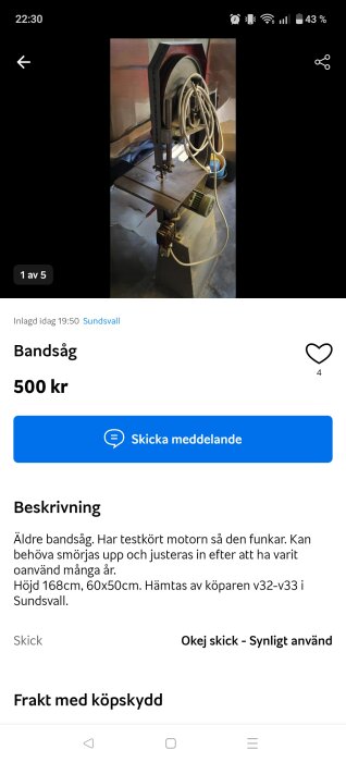 Äldre bandsåg i ett garage, omgiven av elektriska kablar. Sågen står på ett betongfundament och är redo för användning.