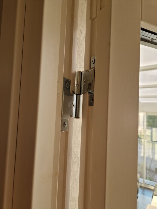 Ett närbild av ett metallgångjärn på en dörr, monterat i en ljus dörrkarm, med skruvar synliga på gångjärnsytorna.