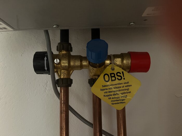 VVS-system med säkerhetsventil och varningsskylt i ett fritidshus, ventiler i svart och blå färg, kopparrör och varningslapp i gult med text.