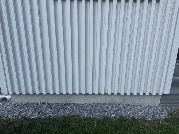 Vit träpanelvägg av ett förråd med betonggrund och grus framför. Gräsmattan sluttar ned mot en vägg.