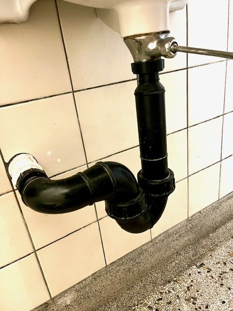 Svart vattenlås under ett handfat monterat utan synliga skarvar framför vitt kakel.