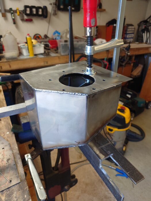 Byggprojekt: En liten tank under konstruktion i verkstaden, fäst med klämma och ser ut att vara av metall. Verktyg och utrustning i bakgrunden.