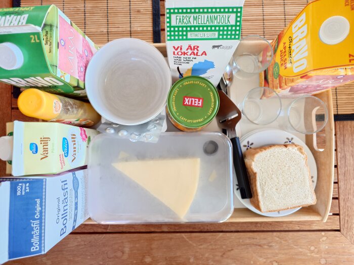 Frukostbricka med mjölk, juice, bröd, ost, krämflaska och glasskålar på ett träbord.
