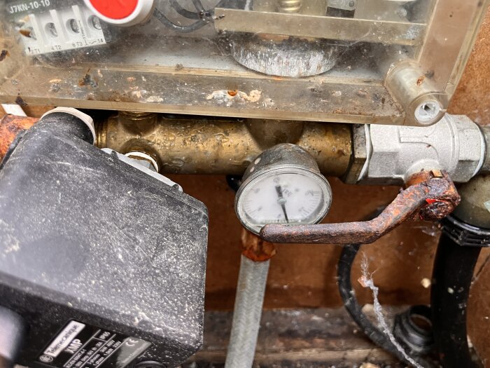 Gammal manometer inbyggd i mässingsrör med rostig ventil och transparent skyddskåpa, visad från en något ovan vinkel.