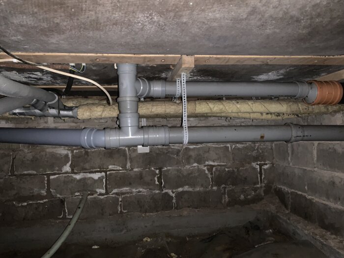 Krypgrund med flera avloppsrör, en orange rörkoppling, och isolerade vattenledningar. Fuktfläckar syns på grundens väggar.
