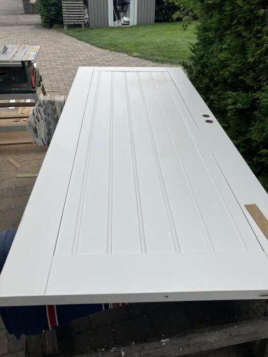 Modifierad vit innerdörr med pärlspontpanel, hål för runt fönster. Dörren ligger på arbetsbockar utomhus, verktyg och verkstad i bakgrunden.