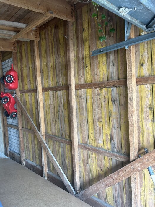 Trävägg i oisolerat garage med panel och bärande bjälkar synliga. Röd leksaksbil hänger på väggen. Byggmaterial ligger på golvet.