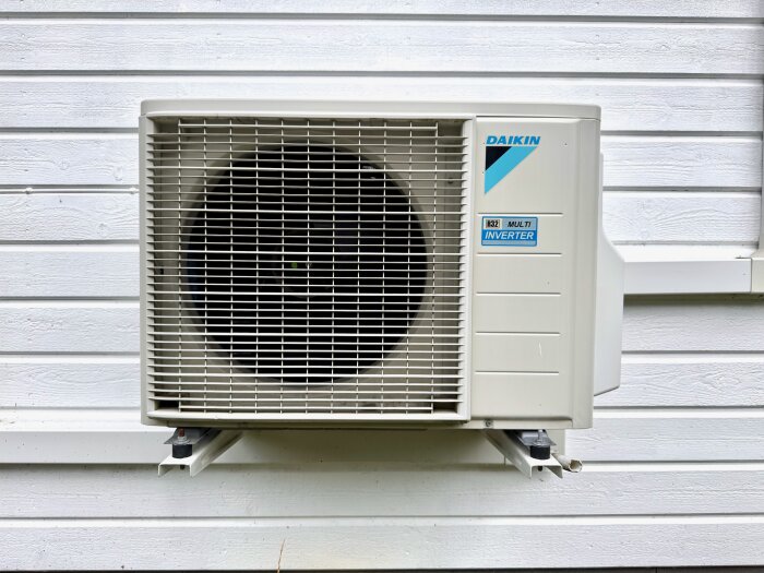 Luftkonditioneringsenhet av märket Daikin, installerad på utsidan av ett vitt trähus, märkt med "R32 Multi Inverter".