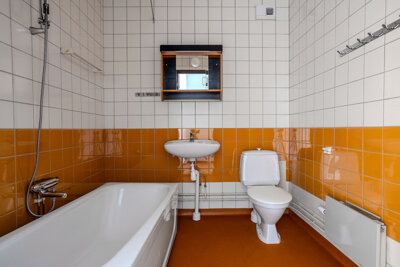 Badrum med badkar, toalettstol, handfat och hylla på vägg. Vitt och orange kakel, tvättställ och fixtur. Brunt golv.