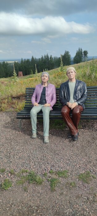 Två realistiska statyer av en sittande kvinna och man på en bänk i en grönskande, kuperad miljö under en molnig himmel.