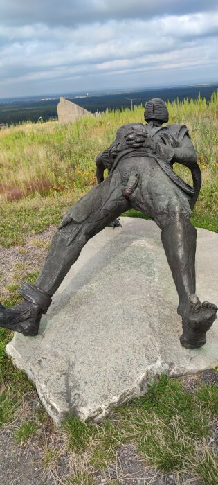 Skulptur av en figursäten som lutar sig bakåt på en klippa, med en expansiv utsikt över landskapet i bakgrunden.