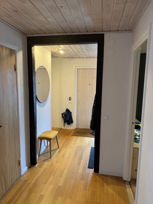 En hall med trägolv och träpaneltak, synlig rund spegel, gul bänk, klädhängare och dörr med ljust trä, delvis synlig svart bjälke.