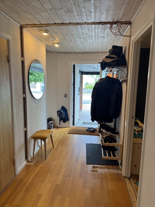 Hall med träpanelstak och trägolv, ett nymonterat skoställ på höger sida, hängande kabeldragningar och en rörig hatthylla.