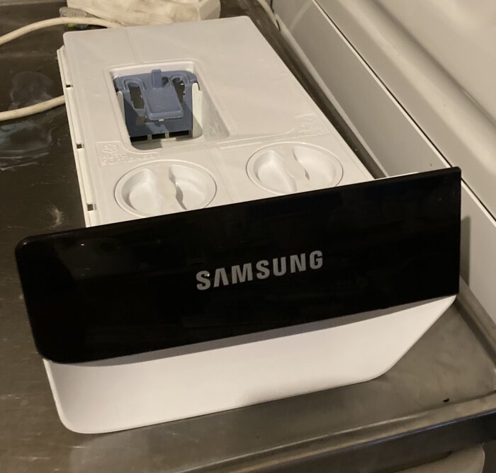 Tvättmedelsskålen i en Samsung tvättmaskin med två runda öppningar markerade i grönt, en med metallspiral, ovanpå en metallisk yta.