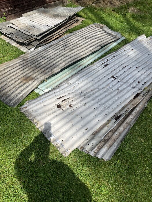 Gamla vågformade takplåtar av metall ligger utspridda på en gräsmatta, med rostfläckar och hål, bredvid ett hus.