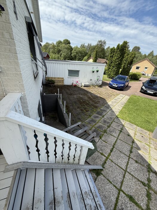 Vit yttervägg, trappa, stenlagd yta och bilar vid en grönskande trädgård; innan renovering.