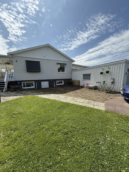 Vit villa med gräsmatta och stensatt gång, blå bil delvis synlig, solig dag med molnig himmel.