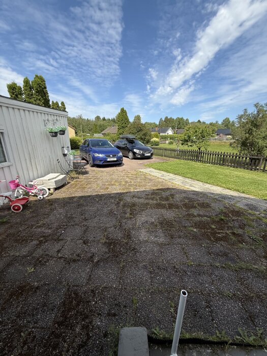 Uppfart med två parkerade bilar bredvid ett vitt hus, en barncykel och en trädgård i bakgrunden under en klarblå himmel.